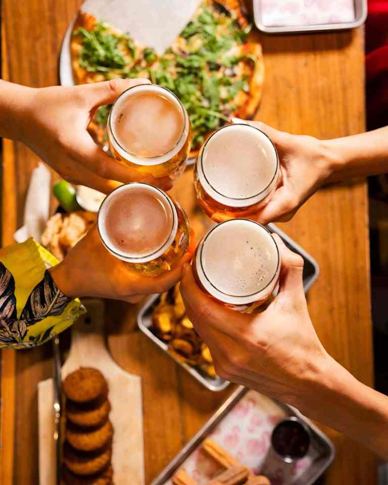 Four hands toasting glasses of craft beer over a table with food at a tasting in Tirana