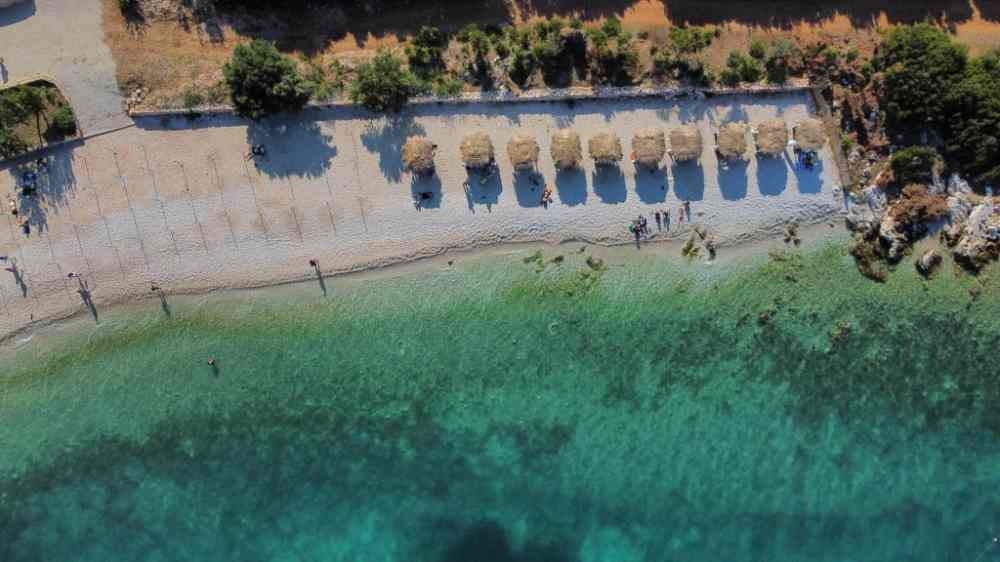 A quiet turquoise bay with sun loungers seen from the water on a Vlora boat tour along the Ionian coast