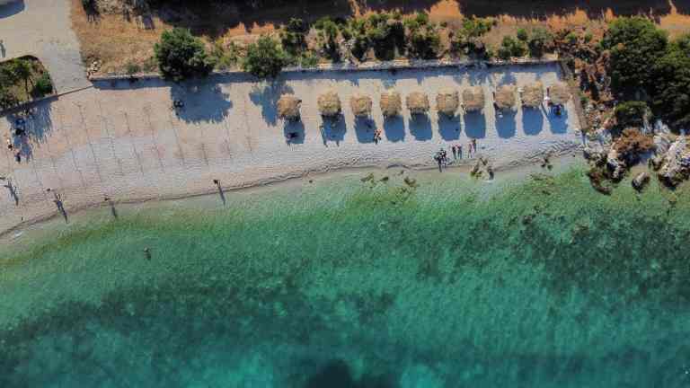 A quiet turquoise bay with sun loungers seen from the water on a Vlora boat tour along the Ionian coast