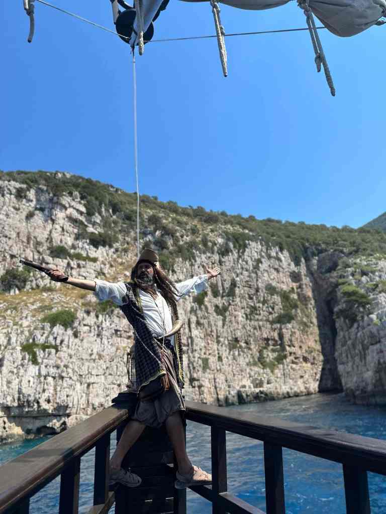 A traveller on the deck of the wooden tour boat with steep Ionian cliffs rising behind on a Vlora boat tour