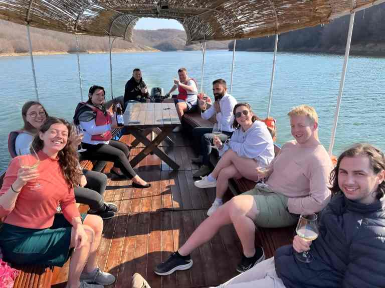 Group of guests sitting at an outdoor table on a deck overlooking water during a winery lunch near Tirana