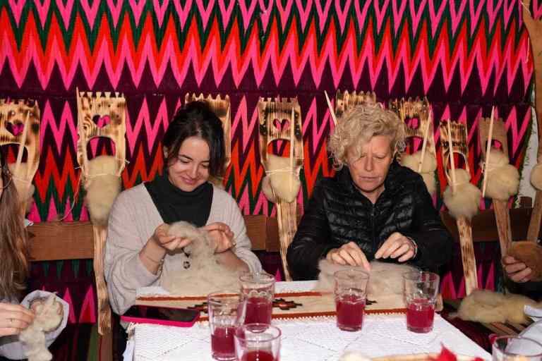 Two women doing traditional handwork in front of a colorful carpet in Lazarat